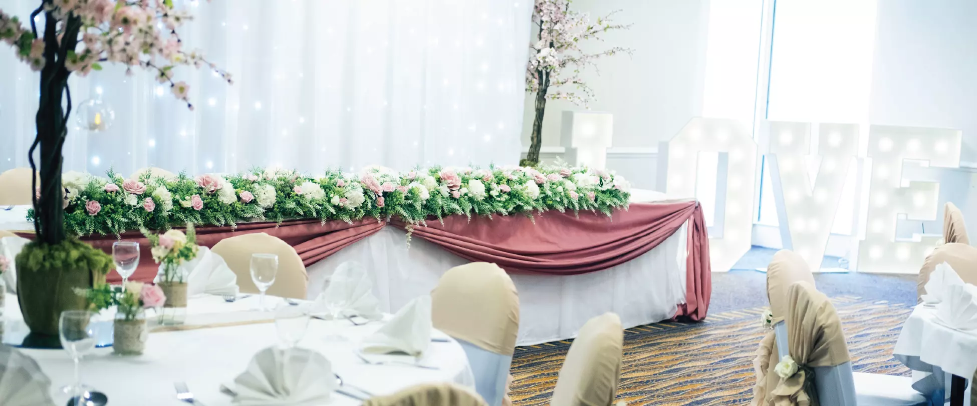 A wedding table set up with a white tablecloth, a pink draped cloth on the from, white and pink flowers, and trees with pink flowers..